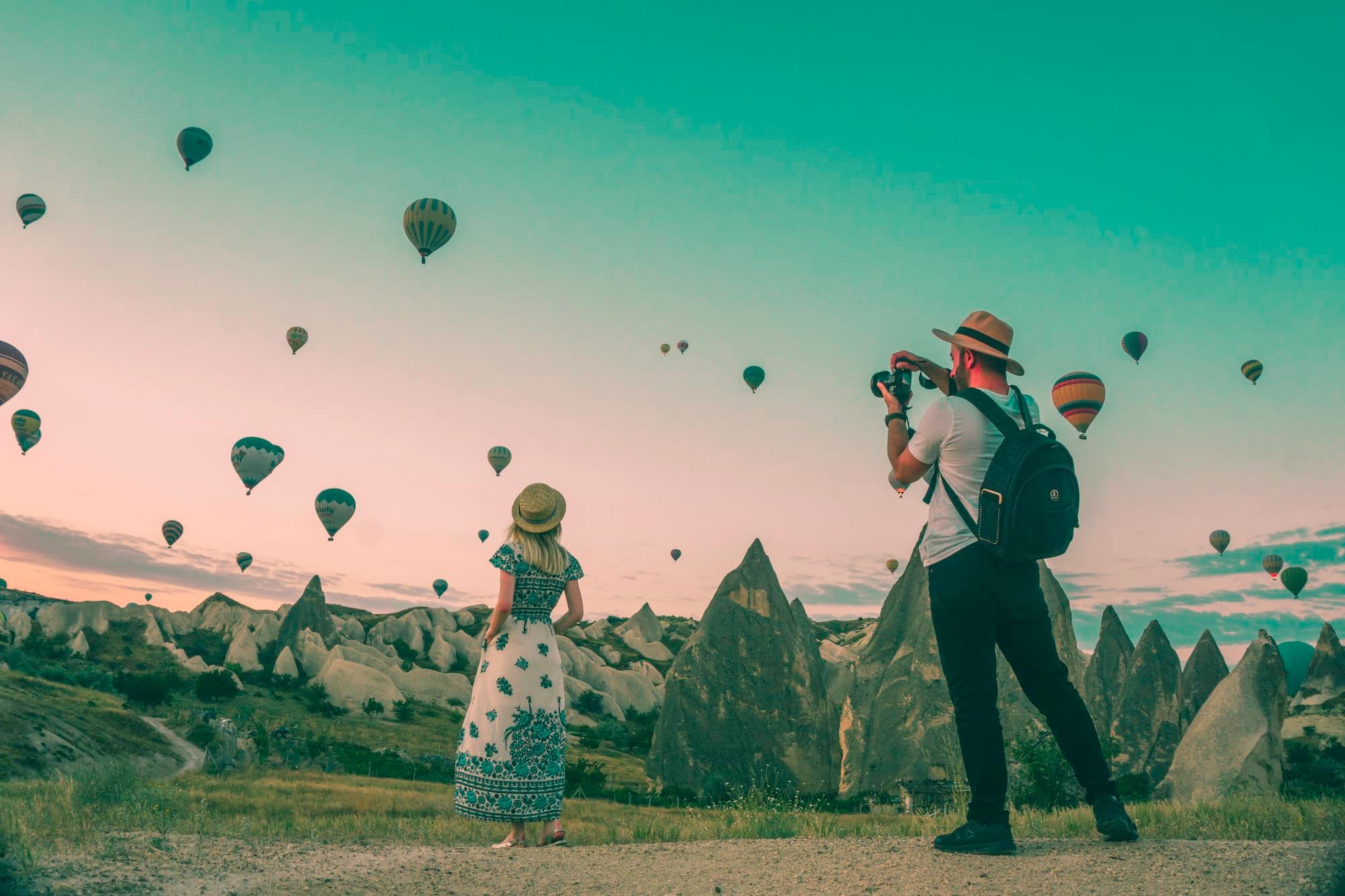 A photographer capturing a traveler at dawn beneath dozens of hot-air balloons in Cappadocia